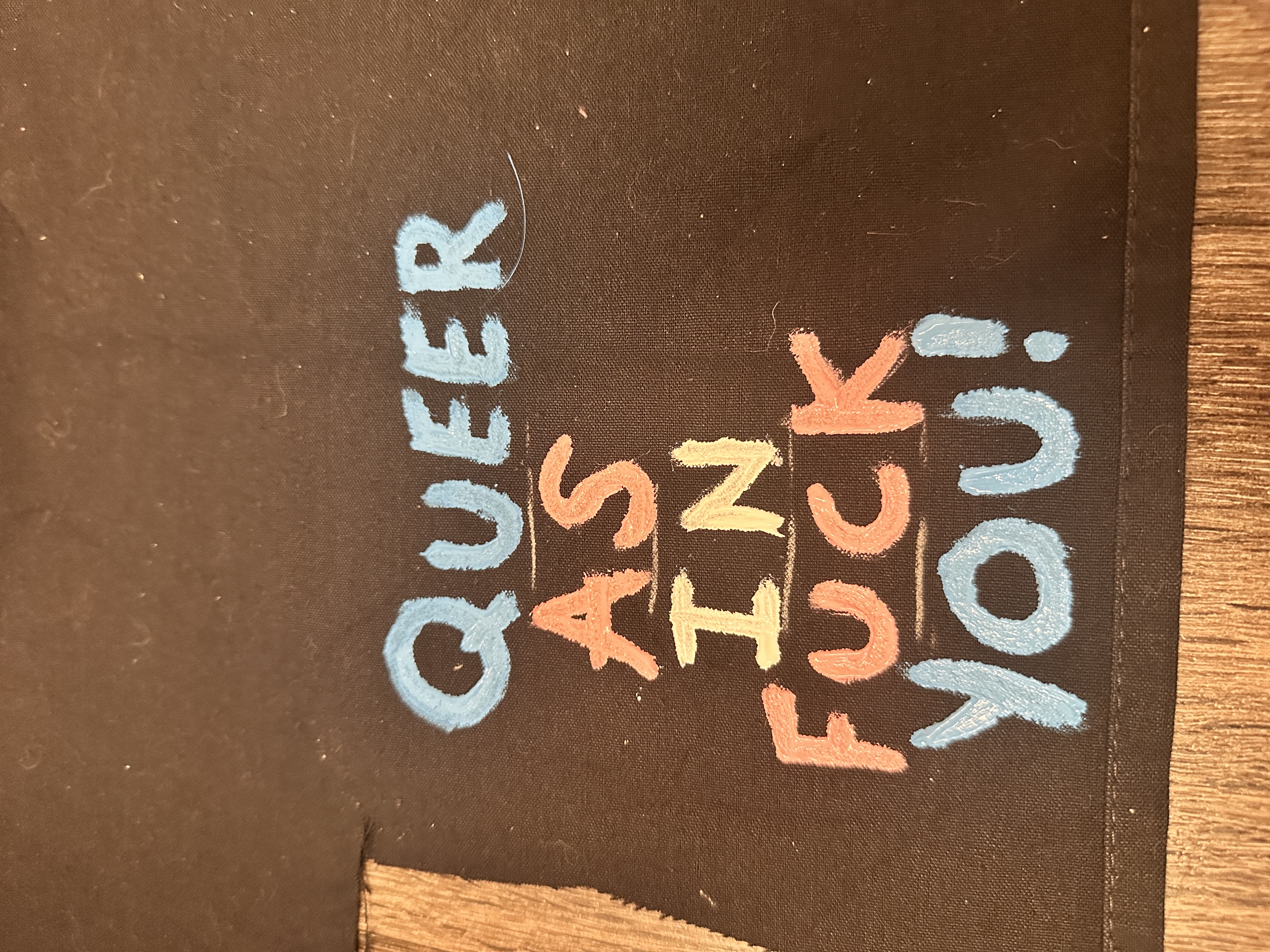 Queer as in Fuck You! painted in trans colors alternating per word on black fabric