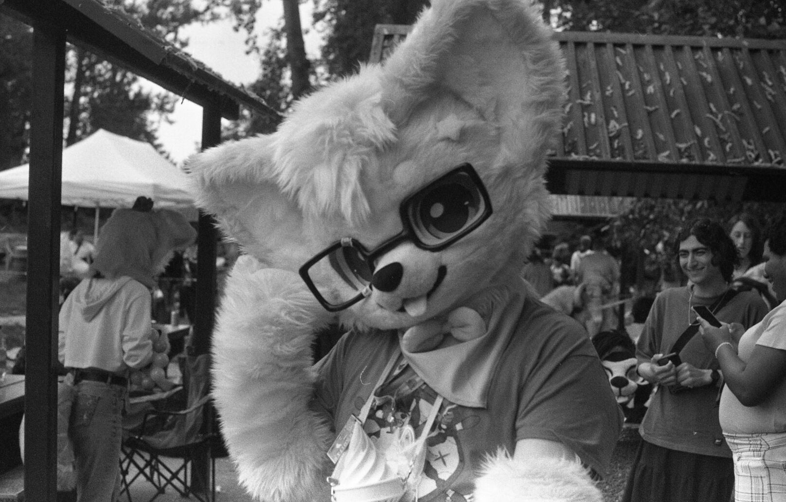 A black and white photo of a partial kemono corgi suiter. There are some other suiters and people behind.